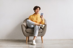 Handsome young Asian guy using cellphone, surfing web or social media, sitting in armchair, enjoying contemporary technologies, checking new mobile app against white studio wall