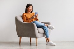 Charming young Asian woman using cellphone, communicating on web, working or learning online, sitting in armchair against white wall, free space. Lovely millennial lady chatting on smartphone