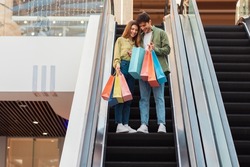 Happy Couple Doing Shopping Standing On Moving Escalator Stairs Holding Shopper Bags In Mall Indoors. Buyers Looking At New Clothes Walking In Hypermarket On Weekend. Sales Season
