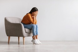 Young Asian woman burying her face in hands and crying, feeling depressed, sitting in armchair against white studio wall, copy space. Millennial lady having mental problems or mood disorder