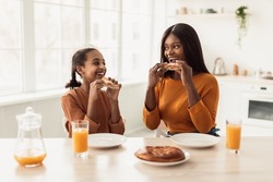 Happy African American Mother And Daughter Having Lunch Eating Sandwiches And Drinking Orange Juice Sitting At Table In Modern Kitchen At Home. Healthy Family Nutrition Concept