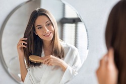 Haircare Concept. Attractive Young Lady Brushing Her Thick Beautiful Hair With Comb While Standing Near Mirror In Bathroom, Happy Woman Wearing White Silk Robe Looking To Her Reflection And Smiling