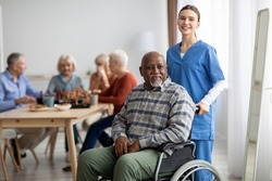 Happy black man older patient on wheelchair with female nurse smiling at camera, group of senior people sitting on couch on background, nursing home interior, healthcare for elderly people concept