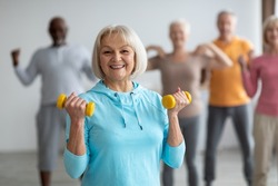 Multiracial group of senior people in sportswear doing strength building fitness exercises with dumbbells, holding fitness tools and smiling at camera, selective focus on positive elderly lady
