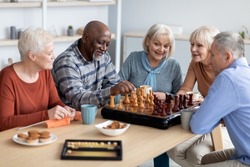 Multiracial senior people men and women in casual playing chess at nursing home, sitting around table, drinking tea with cookies, playing table games, knitting, spending time together