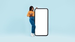 Wow, Great Cool Offer. Full Body Length Of Happy Casual Black Woman Pointing At Big Blank Smartphone Display Showing Huge White Empty Cell Phone Screen Standing On Blue Studio Background Wall, Mock Up