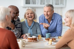 Multiracial group of happy senior people sitting around table drinking tea with cake and having conversation, smiling and laughing, having home party or enjoying time at nursing home