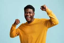 Emotional young black guy in casual raising fists up on blue studio background, celebrating success, happy handsome african american millennial man sharing good emotions, copy space