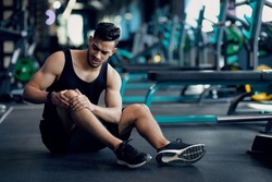 Sport Trauma. Young Arab Male Athlete Touching His Hurted Knee At Gym, Upset Middle Eastern Guy Sitting On Floor And Massaging Sore Injured Leg After Workout Training In Fitness Club, Free Space
