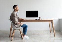 Rear back view of happy Middle Eastern man looking at blank empty computer monitor sitting at table at workplace, watching webinar or having online web video call, free copy space. mockup template