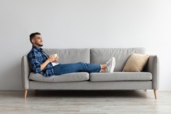 Rest Concept. Happy Arab guy drinking coffee sitting on comfortable couch at home in living room. Cheerful casual man relaxing on sofa, enjoying weekend free time or break from work, full body length