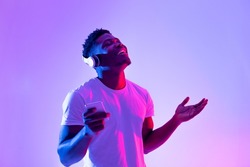 Positive black guy in modern headphones using smartphone, enjoying favorite music with closed eyes in neon light. Young African American man listening to cool playlist or audio book