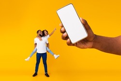 Great Offer. Cheerful black dad carry daughter kid on back and shoulders, excited girl pointing at giant mobile phone showing blank space for mock up, hand holding white empty screen, full body length