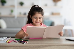 Education Concept. Portrait Of Cute Little Arab Girl Reading Book At Home, Excited Middle Eastern Female Child Study While Sitting At Desk At Home, Smiling Kid Doing Homework, Copy Space
