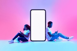Cool mobile app. African American couple sitting near big smartphone with mockup, using modern gadgets, advertising new application or website, promoting your product or service in neon light