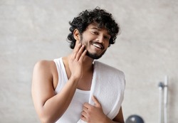 Portrait of happy indian man touching beard and smiling at camera, attractive young guy enjoying morning routine and his appearance. Male facial care