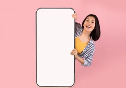 Advertising Concept. Portrait of excited asian lady peeking out big giant vertical smartphone with white blank screen presenting device display. Gadget with empty free space for mock up, pink wall