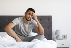 Fatigue Concept. Tired Young Arab Man Sitting In Bed At Home, Exhausted Millennial Middle Eastern Guy Waking Up In Bedroom, Feeling Unwell, Having Headache Or Suffering Insomnia, Free Space
