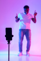 Black male influencer shooting video for social media on mobile phone, having fun in neon light, mockup. African American blogger live streaming content for vlog, broadcasting online, selective focus