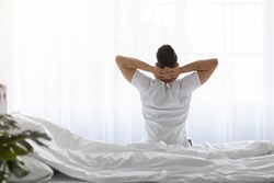 Good Morning. Rear View Of Young Man With Hands Behind Head Sitting On Bed After Waking Up, Unrecognizable Male In White T-Shirt Stretching In Light Bedroom After Awakening, Copy Space