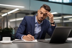 Concentrated exhausted young arab man CEO sitting at worktable in front of computer, taking notes and touching head, looking for creative solutions while crisis, modern office interior, copy space