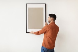 Modern Home Interior And Domestic Decor. Smiling young man hanging painting, putting photo picture frame on the wall. Casual guy holding showing empty poster, design mockup, blank free copy space