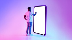 Full length of millennial black guy standing near big cellphone with mockup for your app on screen, interacting with user interface in neon light. Smartphone display template for website or ad