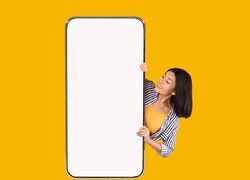 Great Offer. Portrait of excited asian woman peeking out big giant vertical cell phone with white blank screen and looking at device display. Gadget with empty free space mock up, yellow orange wall