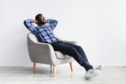 Peaceful young black man relaxing in armchair against white studio wall, free space. Full length of African American guy enjoying lazy weekend morning, resting with hands behind head