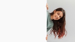 Happy Preteen Girl Posing Hiding Behind Empty White Board And Smiling To Camera In Studio Over Gray Background. Child Holding Paper Poster Advertising Your Text. Panorama, Mockup