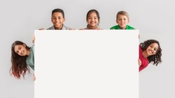 Happy Diverse Kids Hiding Behind Empty White Board Smiling To Camera Posing Over Gray Studio Background. Mixed Boys And Girls Advertising Your Text On Blank Poster. Panorama, Mockup