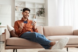Fast Online Shopping. Smiling young Arab guy holding debit credit card in hand and using cell phone, making financial transaction sitting on the couch at home in living room, free copy space