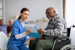 Attractive young woman in blue workwear nurse helping senior black man in wheelchair with questionnaire, african american pensioneer filling papers at nursing home, having assistance