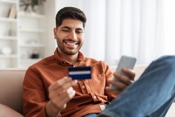 Internet Mobile Shopping Concept. Positive Middle Eastern man using cellphone holding looking at credit card purchasing things online sitting on sofa in living room, buying gifts in web store