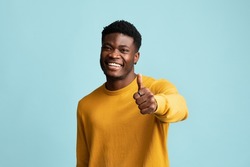 Happy charismatic handsome young black guy showing thumb up and smiling at camera, posing alone on blue studio background, copy space. Positive african american man recommending nice deal