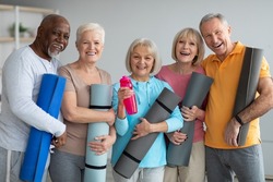 Group of active athletic pensioneers different nationalities in sportrswear attending yoga class at retreat center, holding fitness mats and cheerfully smiling at camera. Sport for senior people