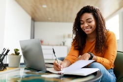 Business And Education Concept. Smiling young black woman sitting at desk working on laptop writing letter in paper notebook, free copy space. Happy millennial female studying using pc