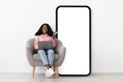 Mobile shopping application. Happy black lady sitting on chair near big cellphone with empty screen using credit card and laptop, white studio wall background, mockup, collage