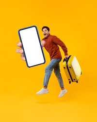 Male Tourist Jumping Holding Huge Smartphone And Suitcase Showing Blank Cellphone Screen To Camera Advertising Great Travel Application Posing In Mid Air On Yellow Background In Studio. Vertical