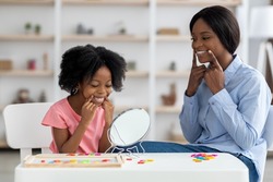 Female speech pathologist young black woman exercising with adorable little girl with language difficulties, working on word sounds, kid looking at mirror, touching mouth and smiling, clinic interior