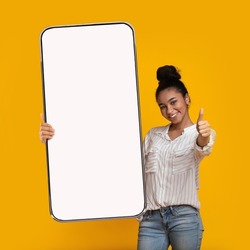 I Recommend. Portrait of smiling black female teenager holding big giant cell phone with white blank device screen in hand, showing thumb up gesture. Gadget with empty free space for mock up, banner