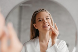 Young woman caring for facial skin using cotton pad and looking in round mirror in bathroom interior. Beauty care and pampering. Daily female skincare routine concept
