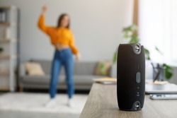 Millennial woman dancing to favorite music at home, selective focus on portable wireless speaker, free space. Young female moving to popular song, using modern stereo system indoors
