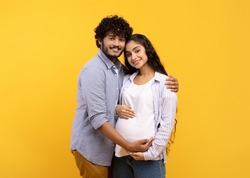 Satisfied young indian man hugging pregnant wife and touching belly, smiling together at camera over yellow background. Couple awaiting baby, cute parents-to-be enjoy tender moment