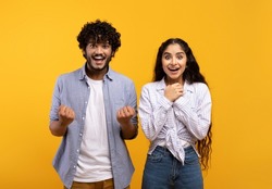 Wow, yes. Portrait of overjoyed indian man and woman cheering and shaking clenched fists, looking at camera in excitement. Happy couple celebrating win over yellow studio background wall
