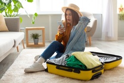 Terrified young woman reading shocking travel news or negative message about tourism on smartphone while packing suitcase at home. Cancelled flight, traveling problems during covid epidemic