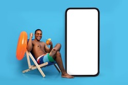 Excited black guy with tropical cocktail chilling in lounge chair near huge smartphone with blank screen for your ad, blue background. man relaxing on beach with cool drink on summer holidays