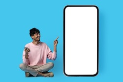 Happy indian guy in casual sitting on floor and pointing at huge smartphone with white blank screen, using mobile phone, blue studio background, full length shot, mockup, panorama with copy space