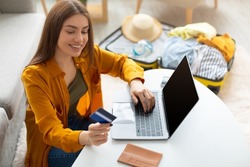 Cheery young lady using credit card and laptop pc with mockup for online travel agency website, booking abroad trip, making hotel reservation on web from home, free space