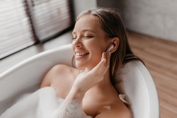 Young lady relaxing in foamy bathtub and listening music in wireless earphones, resting in bathroom at home. Relaxed woman bathing with closed eyes